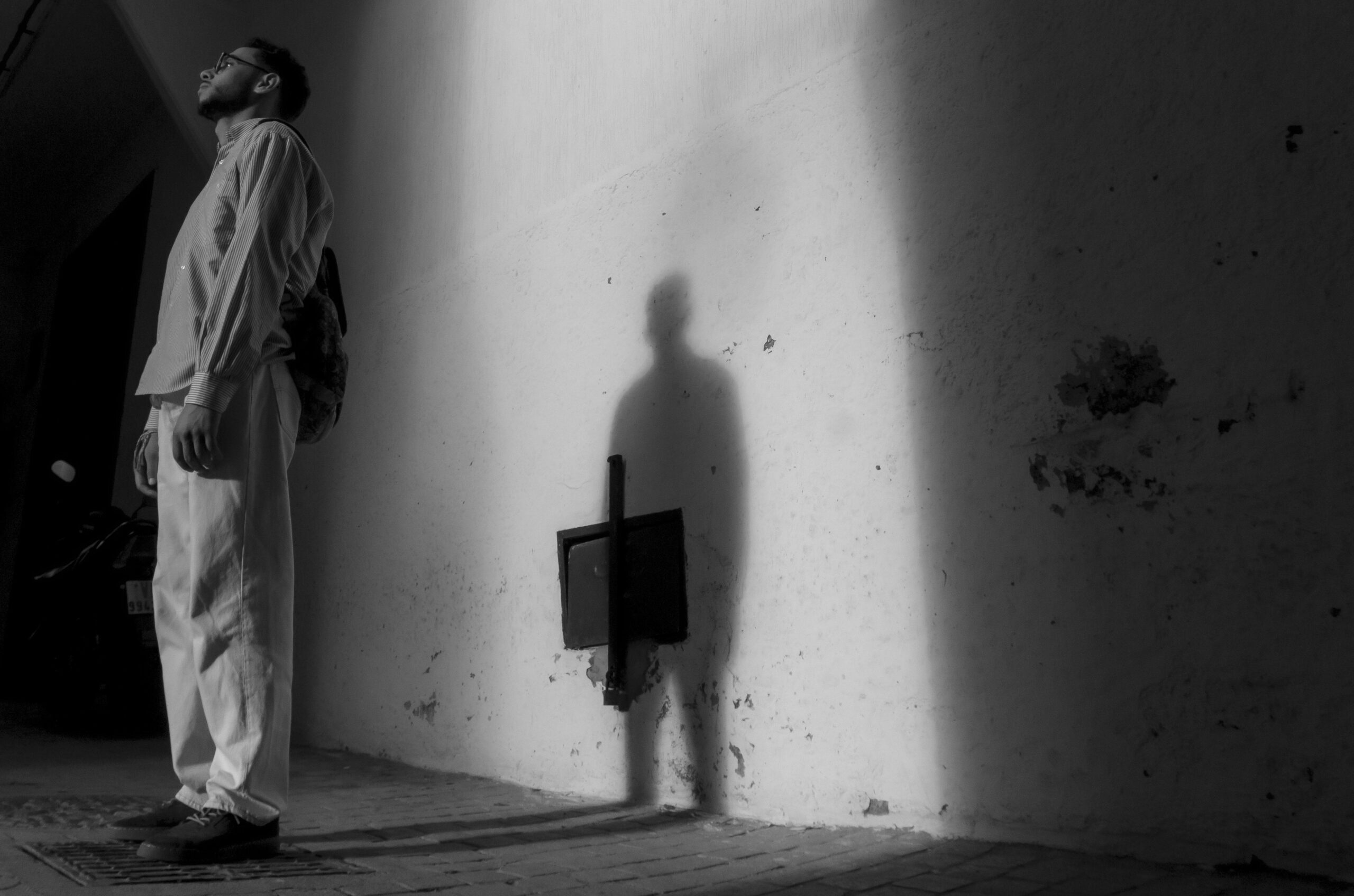 A man in a bright alley stands reflectively with a shadow cast on the wall.